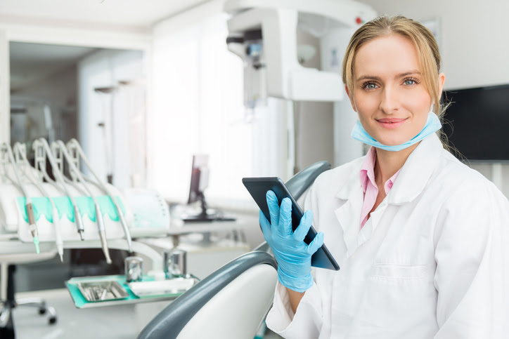 Horizontal color image of female dentist holding digital tablet in dental clinic office, sitting, smiling and looking at camera. Female doctor wearing white uniform, blue surgical mask and gloves. Dental equipment in the background.